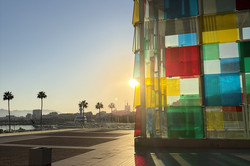 Das Centre Pompidou Málaga bei Sonnenuntergang: Die farbenfrohe Glasfassade fängt das Licht der untergehenden Sonne ein, im Hintergrund die Promenade mit Palmen und dem Hafen.