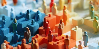 A 3D illustration featuring numerous small, colorful, human-like figures without facial details, standing on cube-shaped blocks of varying heights in shades of blue, orange, yellow, and green. The figures are arranged in groups and vary in size and color. The background is blurred, so the focus is on the figures in the foreground.