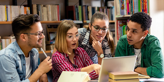 Eine Gruppe Studierender arbeitet gemeinsam in einer Bibliothek. Sie sitzen um einen Tisch mit offenen Büchern und einem Laptop, diskutieren und tauschen sich über ihre Arbeit aus. Im Hintergrund sind Bücherregale mit zahlreichen Fachbüchern zu sehen, was eine konzentrierte Lernatmosphäre vermittelt.
