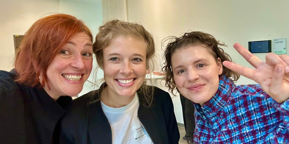 Selfie von Dr. Katty Nöllenburg (links), Dr. Isabell Diekmann von der Universität Paderborn (Mitte) und Leonie Stoll (rechts) auf der DeZiM-Konferenz in Bielefeld. Die drei Frauen stehen eng beieinander und lächeln in die Kamera. Im Hintergrund ist ein heller Konferenzraum mit weißer Wand, Heizkörper und einem kleinen Display an der Wand zu sehen.