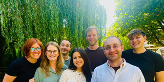 Gruppenfoto von sieben lächelnden Personen im Freien. Die Gruppe steht eng zusammen vor grünen Bäumen und hängenden Weidenzweigen. Im Hintergrund ist helles Sonnenlicht zu sehen, wodurch das Bild sommerlich und freundlich wirkt.