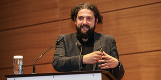 Prof. Dr. Aladin El-Mafaalani stands at a speaker's podium at TU Dortmund University and gives a presentation at the Dortmund Gambrinus Forum. The green “tu” logo and the words “technische universität dortmund” can be seen on the lectern. He is wearing a dark suit and gesturing with his hands. There is a glass of water on the left side of the lectern, and wood paneling is visible in the background.