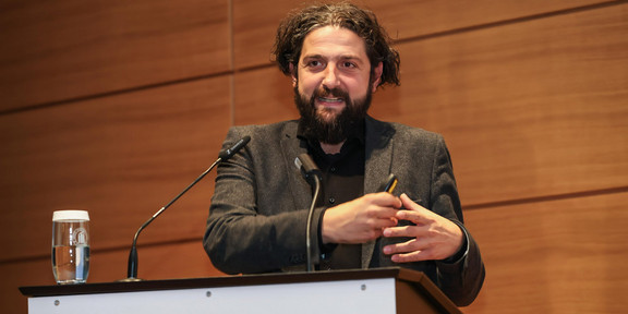 Aladin El-Mafaalani während seines Vortrags am Podium beim Gambrinus-Forum Prof. Dr. Aladin El-Mafaalani steht an einem Rednerpult der Technischen Universität Dortmund und hält eine Präsentation auf dem Dortmunder Gambrinus-Forum. Auf dem Pult ist das grüne „tu“-Logo sowie der Schriftzug „technische universität dortmund“ zu sehen. Er trägt einen dunklen Anzug und gestikuliert mit den Händen. Links auf dem Pult steht ein Glas Wasser, im Hintergrund ist eine Holzvertäfelung sichtbar.