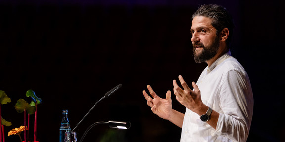 Aladin El-Mafaalani is standing on a stage at a podium, speaking to the audience. He is wearing a white shirt and gesturing with both hands. On the podium are a microphone, a small lamp, a glass, a water bottle, and some papers. Colored flowers can be seen on the left side of the picture; the background is dimly lit.