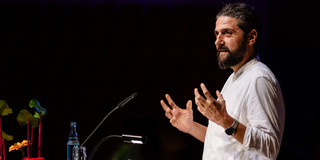 Aladin El-Mafaalani is standing on a stage at a podium, speaking to the audience. He is wearing a white shirt and gesturing with both hands. On the podium are a microphone, a small lamp, a glass, a water bottle, and some papers. Colored flowers can be seen on the left side of the picture; the background is dimly lit.