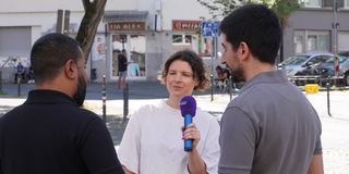Leonie Stoll interviewt mit Mikrofon zwei Männer auf einer belebten Straße in Köln-Kalk, im Hintergrund Geschäfte und Bäume.