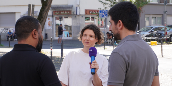 Leonie Stoll interviewt mit Mikrofon zwei Männer auf einer belebten Straße in Köln-Kalk, im Hintergrund Geschäfte und Bäume.