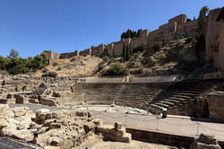 Das römische Theater in Málaga mit seinen steinernen Sitzreihen und einer modernen Holzplattform auf der Bühne. Im Hintergrund erhebt sich die historische Stadtmauer auf einem bewachsenen Hügel unter blauem Himmel.