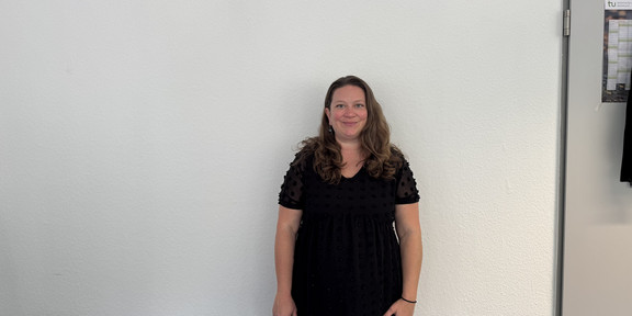 Ganzkörperfoto von Dr. Deborah Warnock mit schulterlangen, welligen Haaren, sie steht vor einer hellen Wand. Sie trägt ein schwarzes Kleid mit kurzen, transparenten Ärmeln und punktförmigem Muster.