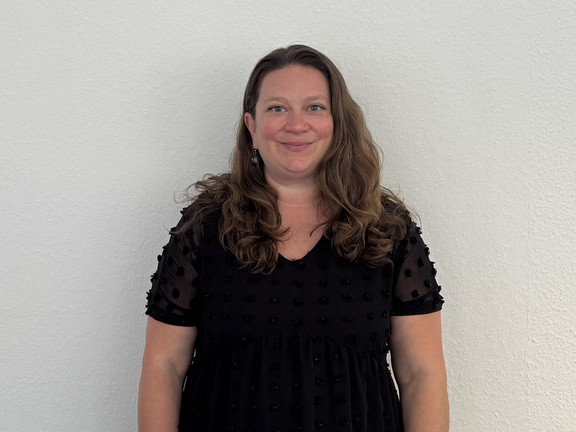 Ganzkörperfoto von Dr. Deborah Warnock mit schulterlangen, welligen Haaren, sie steht vor einer hellen Wand. Sie trägt ein schwarzes Kleid mit kurzen, transparenten Ärmeln und punktförmigem Muster.