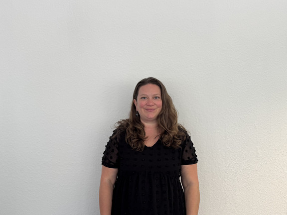 Ganzkörperfoto von Dr. Deborah Warnock mit schulterlangen, welligen Haaren, sie steht vor einer hellen Wand. Sie trägt ein schwarzes Kleid mit kurzen, transparenten Ärmeln und punktförmigem Muster.