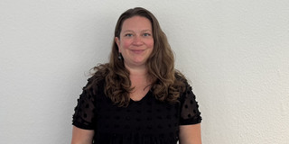 Ganzkörperfoto von Dr. Deborah Warnock mit schulterlangen, welligen Haaren, sie steht vor einer hellen Wand. Sie trägt ein schwarzes Kleid mit kurzen, transparenten Ärmeln und punktförmigem Muster.