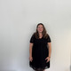 Ganzkörperfoto von Dr. Deborah Warnock mit schulterlangen, welligen Haaren, sie steht vor einer hellen Wand. Sie trägt ein schwarzes Kleid mit kurzen, transparenten Ärmeln und punktförmigem Muster.