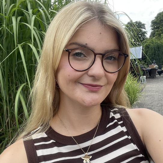 Porträtfoto von Lia-Marie Seil. Sie trägt ein ärmelloses, braun-weiß gestreiftes Oberteil und eine Halskette. Im Hintergrund sind hohe grüne Pflanzen, eine Terrasse und Gartenmöbel zu sehen.