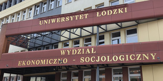 Fassade eines mehrstöckigen Gebäudes der Universität Łódź (Polen) mit gelben und grauen Wandflächen sowie zahlreichen Fenstern. Zwei maroon-rote Vordächer mit goldenen Aufschriften überspannen den Eingangsbereich: Das obere trägt den Schriftzug „UNIWERSYTET ŁÓDZKI", das untere „WYDZIAŁ EKONOMICZNO-SOCJOLOGICZNY" (Fakultät für Wirtschaft und Soziologie). Unter dem unteren Vordach ist ein Eingang mit Glasfront und dem Buchstaben „A" zu sehen, daneben weitere Fenster. Im Hintergrund ist ein Teil eines Backsteinbaus erkennbar.