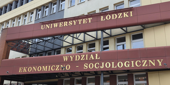 Fassade eines mehrstöckigen Gebäudes der Universität Łódź (Polen) mit gelben und grauen Wandflächen sowie zahlreichen Fenstern. Zwei maroon-rote Vordächer mit goldenen Aufschriften überspannen den Eingangsbereich: Das obere trägt den Schriftzug „UNIWERSYTET ŁÓDZKI", das untere „WYDZIAŁ EKONOMICZNO-SOCJOLOGICZNY" (Fakultät für Wirtschaft und Soziologie). Unter dem unteren Vordach ist ein Eingang mit Glasfront und dem Buchstaben „A" zu sehen, daneben weitere Fenster. Im Hintergrund ist ein Teil eines Backsteinbaus erkennbar.