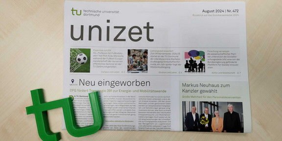 Eine Ausgabe der Hochschulzeitung "unizet" der Technischen Universität Dortmund liegt auf einem hellen Holztisch. Die Titelseite trägt die Überschrift „Neu eingeworben“ und berichtet über die Förderung des Transregio 391 zur Energie- und Mobilitätswende durch die DFG. Weitere Artikel behandeln Themen wie Fußball-Expertise zur Europameisterschaft, Lehrangebote im Wintersemester und Forschungspartnerschaften. Links unten liegt ein grünes plastisches TU-Logo neben der Zeitung. Die Ausgabe ist mit „August 2024 | Nr. 472“ datiert.