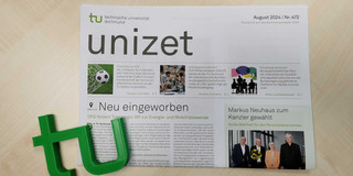 unizet August 2024 Eine Ausgabe der Hochschulzeitung "unizet" der Technischen Universität Dortmund liegt auf einem hellen Holztisch. Die Titelseite trägt die Überschrift „Neu eingeworben“ und berichtet über die Förderung des Transregio 391 zur Energie- und Mobilitätswende durch die DFG. Weitere Artikel behandeln Themen wie Fußball-Expertise zur Europameisterschaft, Lehrangebote im Wintersemester und Forschungspartnerschaften. Links unten liegt ein grünes plastisches TU-Logo neben der Zeitung. Die Ausgabe ist mit „August 2024 | Nr. 472“ datiert.