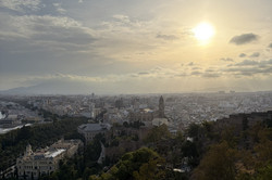 Panoramaansicht der Stadt Málaga vom Hügel des Castillo de Gibralfaro aus, aufgenommen bei Sonnenuntergang. Die Sonne bricht durch eine Wolkendecke und wirft warmes Licht über die dichte Bebauung der Stadt, in deren Zentrum der Turm der Kathedrale von Málaga herausragt. Im Vordergrund erstreckt sich ein bewachsener Hang mit Bäumen, Sträuchern und Agaven, links unten ist ein großes historisches Gebäude mit weißer Fassade zu sehen. Am Horizont sind Berge und links das Meer zu erkennen.