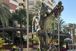 Die Bronzestatue der Medusa in Marbella auf einem steinernen Sockel, im Hintergrund Palmen, Blumenbeete und Wohnhochhäuser am Platz.