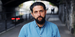 Prof. Dr. Aladin El-Mafaalani is standing on a sidewalk under a bridge. In the background, you can see a street with cars driving by and two pedestrians. The person is wearing a blue denim shirt, and the surroundings appear urban with stone walls and railings along the path.