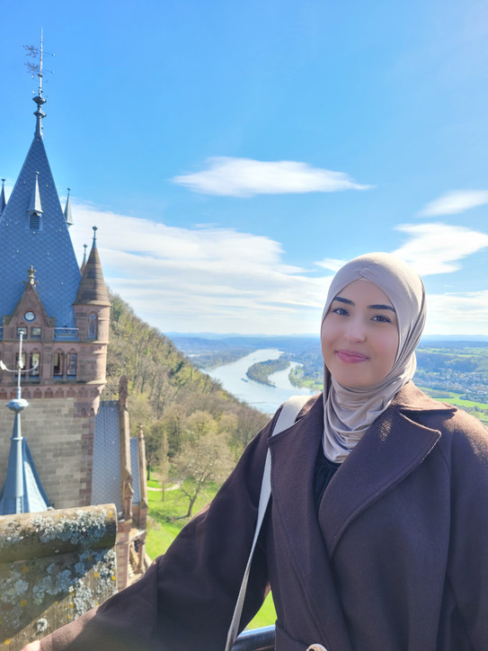 Porträtfoto von Bezanur Bayrak mit hellem Kopftuch und dunklem Mantel auf einer Aussichtsplattform. Im Hintergrund sind ein historisches Schlossgebäude mit Turm, eine Flusslandschaft, grüne Hügel und ein weiter blauer Himmel mit Wolken zu sehen.