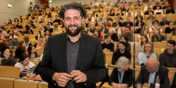 Prof. Dr. Aladin El-Mafaalani steht in einem großen Hörsaal vor einem Publikum. Die Sitzreihen sind mit zahlreichen Zuhörenden besetzt, die aufmerksam einer Veranstaltung folgen. Er trägt einen dunklen Anzug und steht im Vordergrund, während der Hintergrund den vollen Saal zeigt.