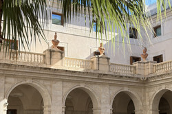 Innenhof (Patio) des Museo de Málaga mit klassizistischer Architektur: Eine Reihe weißer Steinbögen mit Balustraden zieht sich über zwei Etagen, auf den Sockeln zwischen den Bögen stehen drei marmorne Büsten. Im Vordergrund hängen grüne Palmenblätter von oben ins Bild, darüber ist ein Glasdach mit Metallkonstruktion zu erkennen, durch das Tageslicht einfällt. Die Wände sind hell verputzt, dunkle Holztüren und Fenster rahmen die Bögen ab.
