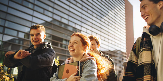 Eine Gruppe junger Menschen steht vor einem modernen Gebäude mit Glasfassade. Sie tragen lässige Kleidung und halten Bücher und Notizblöcke in den Händen, was auf eine studentische Umgebung hinweist. Die Sonne scheint und erzeugt eine warme, lebendige Atmosphäre.