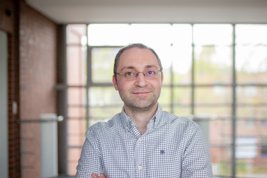 Porträtfoto von Dr. Fatih Kaya mit verschränkten Armen in einem Innenraum mit großen Fensterflächen im Hintergrund. Er trägt ein kariertes Hemd. Hinter ihm sind eine Glasfront und ein heller Flurbereich zu sehen.