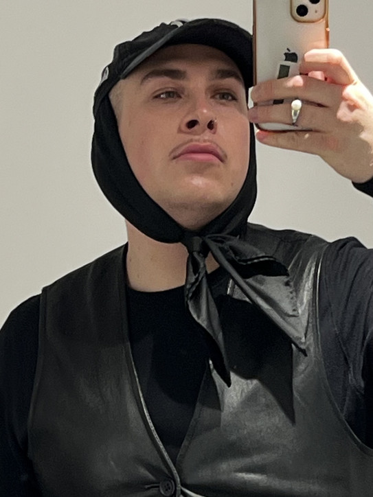 Porträtfoto von Kevin Sikeew, ein Spiegelselfie von einem Smartphone. Er trägt eine schwarze Kopfbedeckung, die unter dem Kinn gebunden ist, sowie ein schwarzes Oberteil mit einer ärmellosen, lederähnlichen Weste. Der Hintergrund ist schlicht und hell.