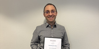 Dr. Fatih Kaya stands in front of a light-colored wall, holding a framed certificate titled “Avicenna Promotionspreis 2025” in his hands. The scene appears formal and depicts the awarding of an academic achievement.