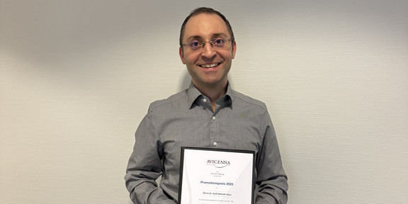 Dr. Fatih Kaya stands in front of a light-colored wall, holding a framed certificate titled “Avicenna Promotionspreis 2025” in his hands. The scene appears formal and depicts the awarding of an academic achievement.