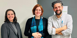 Drei Personen (siehe Bildunterschrift) stehen vor einer grauen Tür und einer hellen Wand. Die Person links trägt einen grauen Blazer und Pullover, die mittlere Person ein schwarzes Outfit mit einem blau-türkisen Schal, und die Person rechts ein hellgraues Hemd mit einem blauen Stift in der Brusttasche. Alle stehen nebeneinander und lächeln in Richtung Kamera.