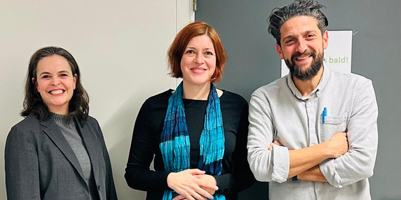 Drei Personen (siehe Bildunterschrift) stehen vor einer grauen Tür und einer hellen Wand. Die Person links trägt einen grauen Blazer und Pullover, die mittlere Person ein schwarzes Outfit mit einem blau-türkisen Schal, und die Person rechts ein hellgraues Hemd mit einem blauen Stift in der Brusttasche. Alle stehen nebeneinander und lächeln in Richtung Kamera.