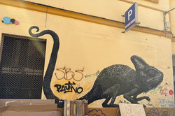 Großes Wandbild (Mural) im SoHo-Viertel von Málaga: Ein schwarzes Chamäleon mit langem, eingerolltem Schwanz, detailliert in feinen Punktstrukturen auf einer gelben Gebäudefassade gemalt. Auf der Wand sind zudem mehrere Graffiti-Tags und Kritzeleien in Schwarz und Farbe zu erkennen. Oben rechts hängt ein blaues Parkhaus-Schild mit dem Buchstaben „P" und dem Schriftzug „APARCAMIENTOS", oben links sind bunte Kreise als Dekoration angebracht. Links befindet sich ein Fenster mit Metallgitter. Im Vordergrund stehen eine Holzbank, Holzpalletten, Pappe und ein aufgestelltes Holzbrett auf dem gepflasterten Boden.