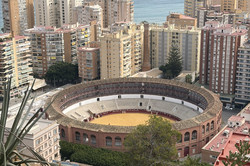 Blick von oben auf die runde Stierkampfarena (Plaza de Toros) von Málaga mit ihrem sandfarbenen Innenraum und den umlaufenden Tribünen. Die Arena ist von mehrstöckigen Wohngebäuden umgeben, dahinter erstreckt sich das Meer mit mehreren Segelbooten. Im Vordergrund sind grüne Pflanzen und ein Holzgeländer auf einem bewachsenen Hang zu sehen.