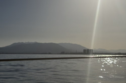 Blick vom Boot auf eine ruhige Wasserfläche mit funkelnden Sonnenreflexen. Rechts oben steht die Sonne tief am Himmel und wirft einen hellen Lichtstrahl ins Bild. Am Horizont erhebt sich eine Bergkette im Dunst, davor sind Silhouetten von Gebäuden zu erkennen. Unten rechts ist die metallene Kante des Boots sichtbar.