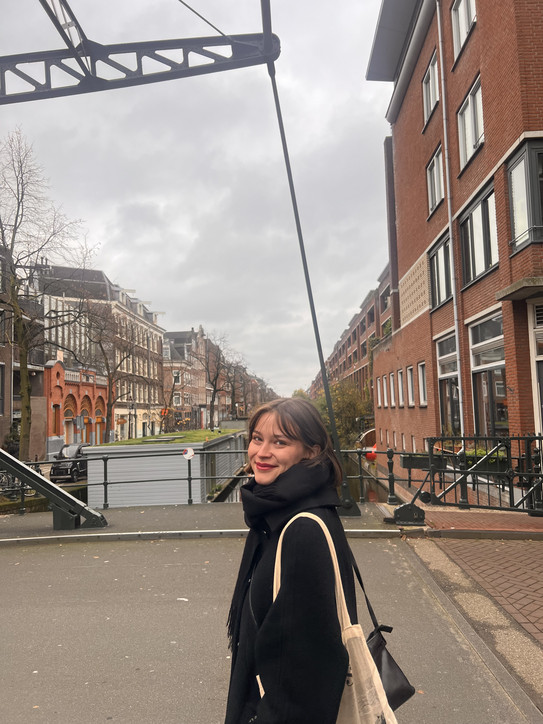 Porträtfoto von Jolina Schyma auf einer Brücke in einer städtischen Umgebung. Sie trägt einen schwarzen Mantel und Schal sowie eine helle Tasche über der Schulter und blickt lächelnd in die Kamera. Im Hintergrund sind ein Kanal, Backsteingebäude, Straßen und ein bewölkter Himmel zu sehen.