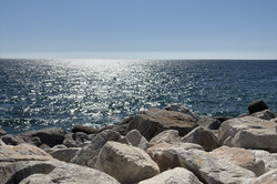 Blick auf die Küste von Marbella an der Costa del Sol: Im Vordergrund füllen große, hellgraue bis beige Felsbrocken (Rippen) den unteren Bildbereich, ihre raue, von der Sonne beschienene Oberfläche ist deutlich zu erkennen. Dahinter erstreckt sich das tiefblaue Meer bis zum flachen Horizont, auf dem die Sonne in zahlreichen hellen Reflexen glitzert. Darüber liegt ein wolkenloser, klarer blauer Himmel.