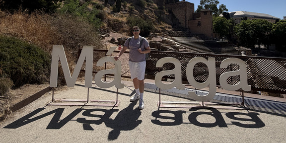 Dustin Cirkel stands smiling in front of the large white letters spelling out “Málaga” and poses for the photo. In the background, the stone rows of seats of the Roman theater and the historic city wall on the hill can be seen against a blue sky.