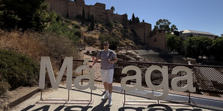 Dustin Cirkel stands smiling in front of the large white letters spelling out “Málaga” and poses for the photo. In the background, the stone rows of seats of the Roman theater and the historic city wall on the hill can be seen against a blue sky.
