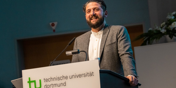 Prof. Dr. Aladin El-Mafaalani steht an einem Rednerpult der Technischen Universität Dortmund und spricht in einem Hörsaal. Auf dem Pult ist das Logo der TU Dortmund mit grünem „tu“-Symbol und Schriftzug zu sehen. Im Hintergrund sind eine Tür und Blumenschmuck sichtbar.
