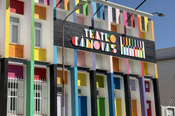 Die farbenfrohe Fassade des Teatro Cánovas in Málaga, gestaltet in leuchtenden vertikalen Streifen und geometrischen Formen in Rot, Gelb, Grün, Blau, Pink und Orange auf weißem Grund. In der Mitte prangt auf einem schwarzen Banner der Schriftzug „TEATRO CÁNOVAS" in bunten Buchstaben, daneben ein farbiges Streifenmuster. Im Vordergrund sind eine Straßenlaterne und ein rotes Auto zu sehen, oben ein klarer blauer Himmel mit mehreren Straßenlaternen.