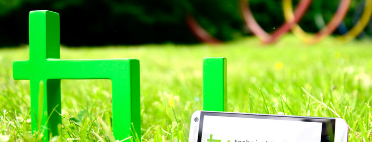 The green logo of TU Dortmund University is standing on a meadow. A smartphone is leaning against it.