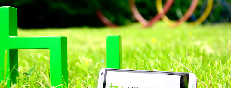 TU-App Ein grünes TU-Logo steht auf grünem Rasen. Daran lehnt ein Smartphone.